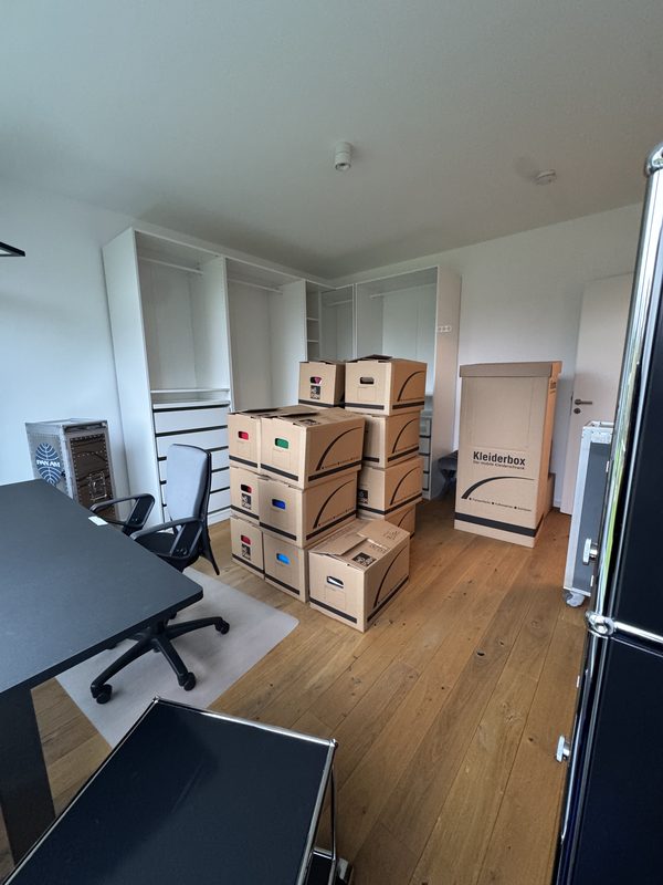 Ein Arbeitszimmer mit schwarzem Schreibtisch, Bürostuhl und einem Stapel beschrifteter Umzugskartons vor einem weißen Einbauschrank