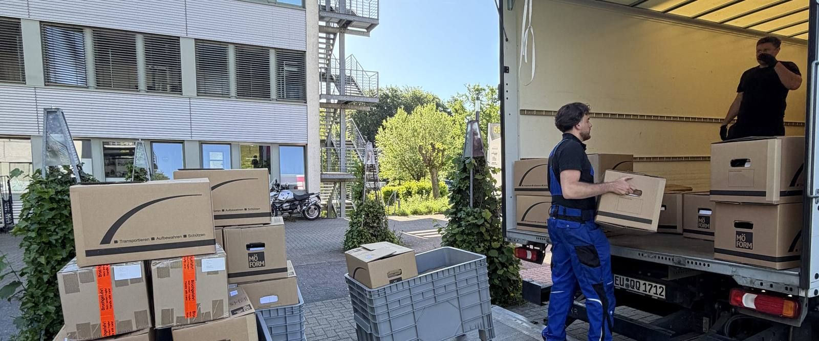 Einblicke in unsere Arbeit in Köln: Vor einem Bürogebäude mit Außentreppe steht ein Umzugswagen mit offenem Laderaum, ein Mitarbeiter trägt einen Karton zur Ladebordwand, ein zweiter steht im Laderaum