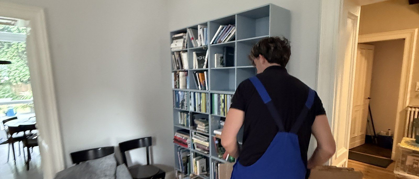 Häufige Fragen zum Umzug in Köln: Ein Mitarbeiter in Latzhose packt Bücher und Ordner aus einem hellgrauen Würfelregal in einen Karton, davor ein Esstisch mit Stühlen