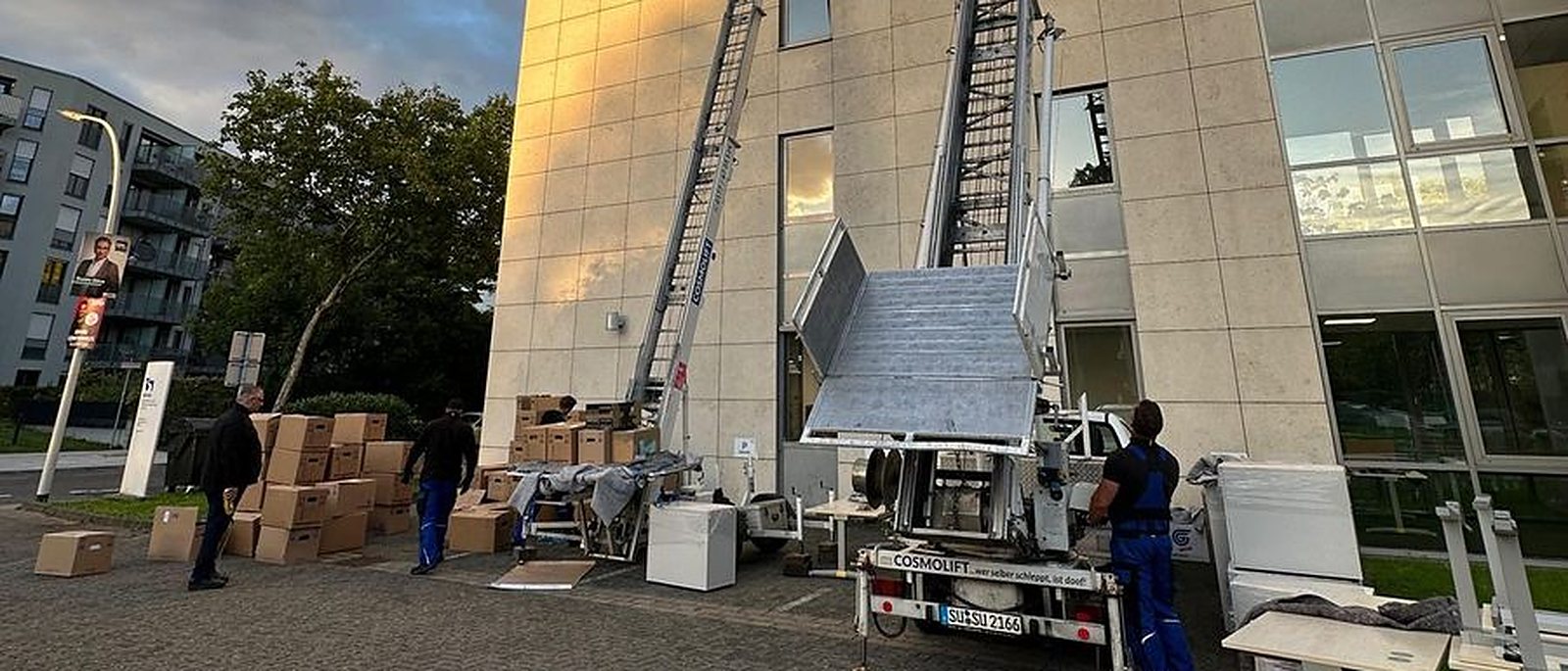 Firmenumzug in Köln: Zwei Möbellifte an der Fassade eines mehrstöckigen Bürogebäudes, davor gestapelte Kartons und Büromöbel auf dem Hof