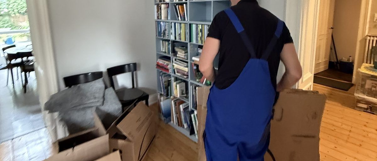 Haushaltsauflösung in Köln: Ein Mitarbeiter in Latzhose trägt einen gefalteten Karton an einem vollen Bücherregal vorbei, davor Stühle und Kartons