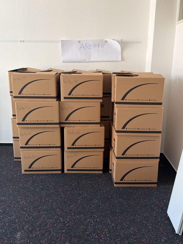 Nummerierte Umzugskartons stapeln sich unter einem handgeschriebenen Schild in einem leeren Büroraum