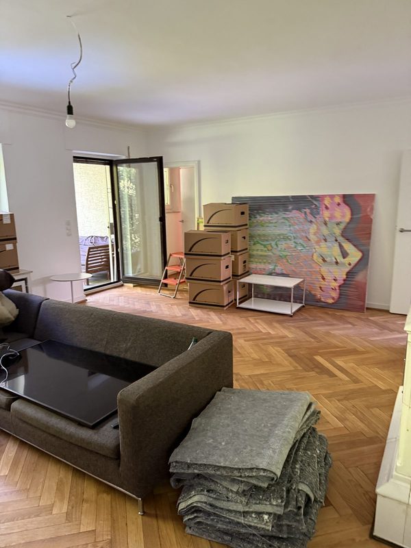 Wohnzimmer mit gestapelten Kartons an der Wand und zusammengelegten Umzugsdecken