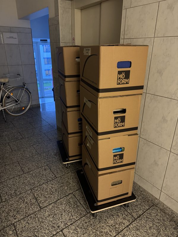 Vier Umzugskartons übereinander auf einem Rollbrett in einem gefliesten Hausflur, daneben ein abgestelltes Fahrrad