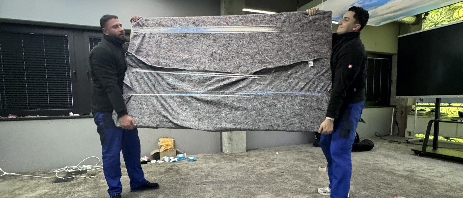 Kontakt zur Umzugsunion in Köln: Zwei Mitarbeiter tragen ein großes flaches Möbelstück in einer grauen Umzugsdecke über einen Hof an einer Hauswand entlang