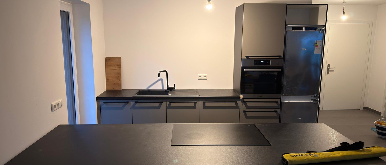 Küchenmontage in Köln: Eine fertig montierte Küche mit dunklen Fronten, Kochinsel mit Induktionsfeld, Spüle, Backofen und Kühlschrank in einem hellen leeren Raum