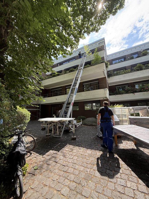 Möbellift steht ausgefahren an einem mehrgeschossigen Wohnhaus, ein Mitarbeiter wartet unten