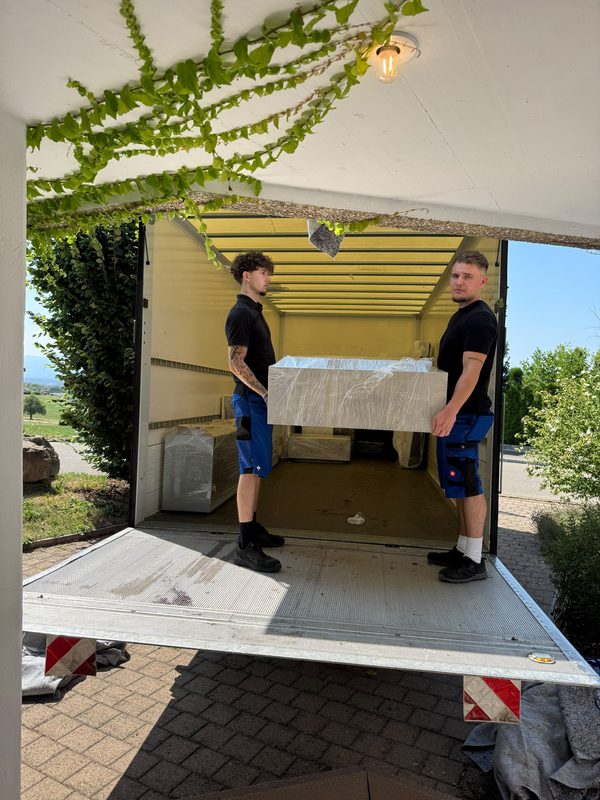 Zwei Umzugshelfer tragen ein foliertes helles Möbelstück über die Ladebordwand in den offenen Kofferwagen, über dem Carport ranken Efeublätter