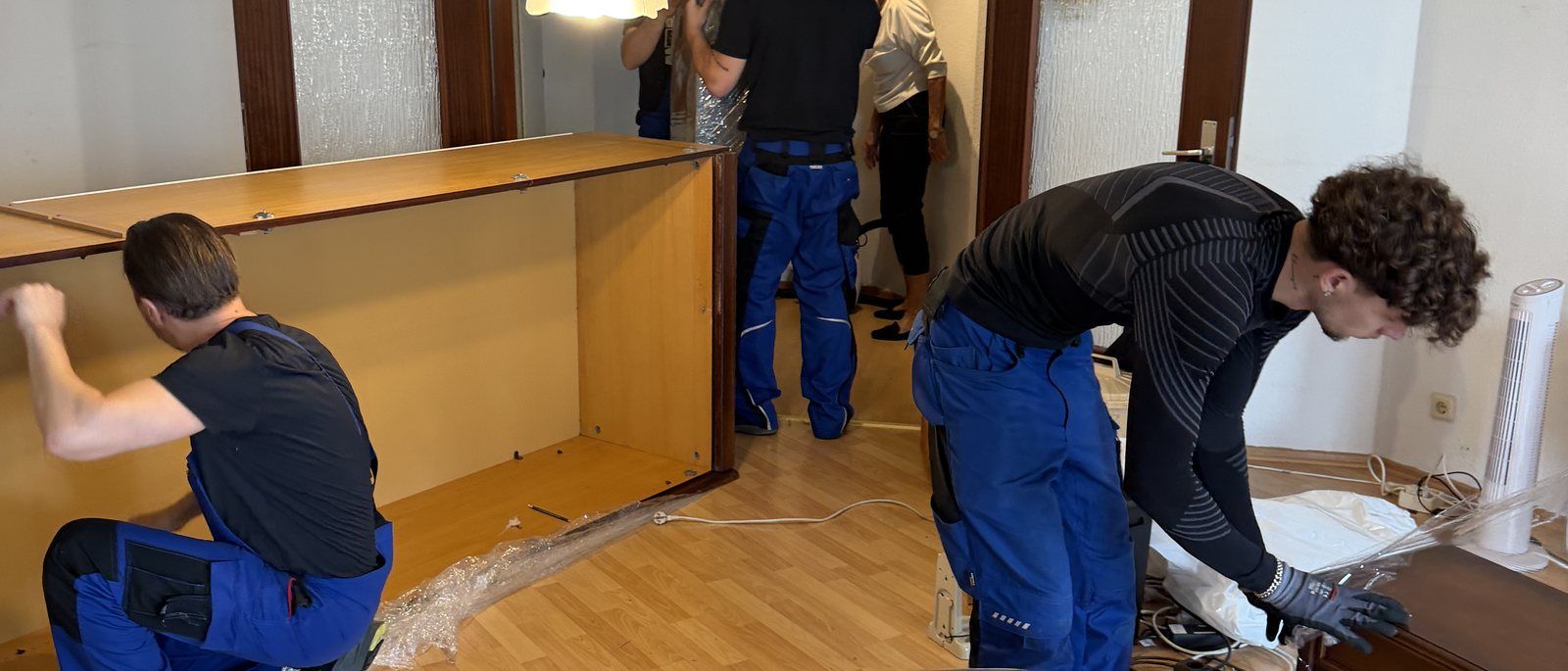 Montageservice in Köln: Drei Mitarbeiter arbeiten in einem Wohnzimmer an einer zerlegten Schrankwand aus Holz, einer hält ein Seitenteil, ein anderer beugt sich über Folie und Schrauben