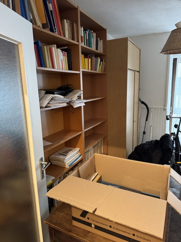 Ein Holzregal mit Büchern und losen Papieren, davor ein geöffneter leerer Umzugskarton auf einem Tisch, bei einem Umzug in Köln