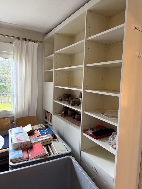 Ein weißes Regalsystem ist bis auf ein paar Bücher und eine graue Kiste im unteren Fach leergeräumt, an der Wand steht handschriftlich WZ
