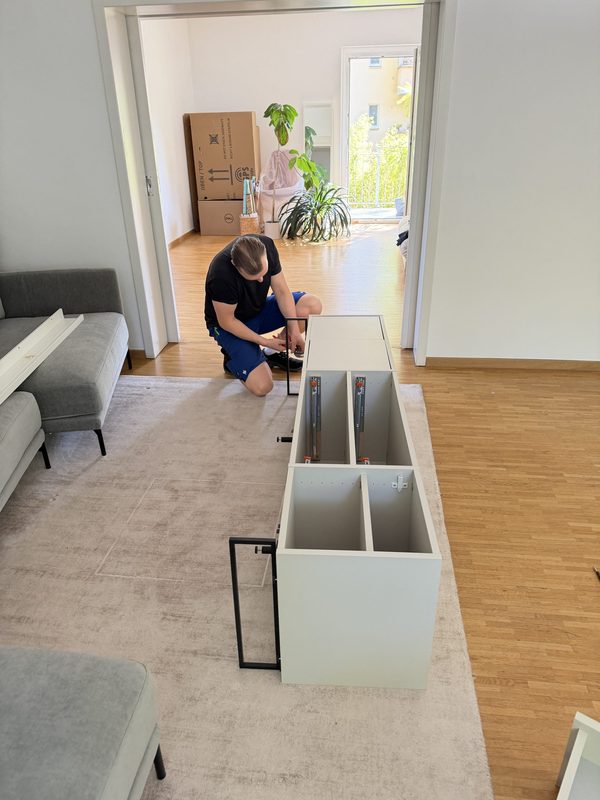 Ein Umzugshelfer kniet auf einem hellen Teppich und montiert ein weißes Sideboard, dessen Korpus noch auf dem Boden liegt, daneben steht das Metallgestell