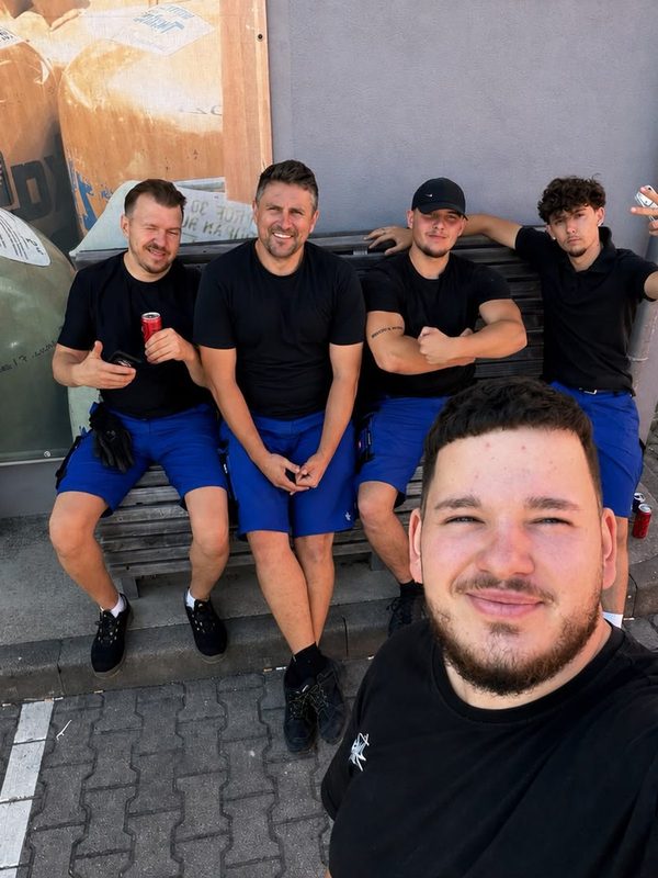 Fünf Mitarbeiter in Arbeitskleidung sitzen zusammen auf einer Bank in der Sonne