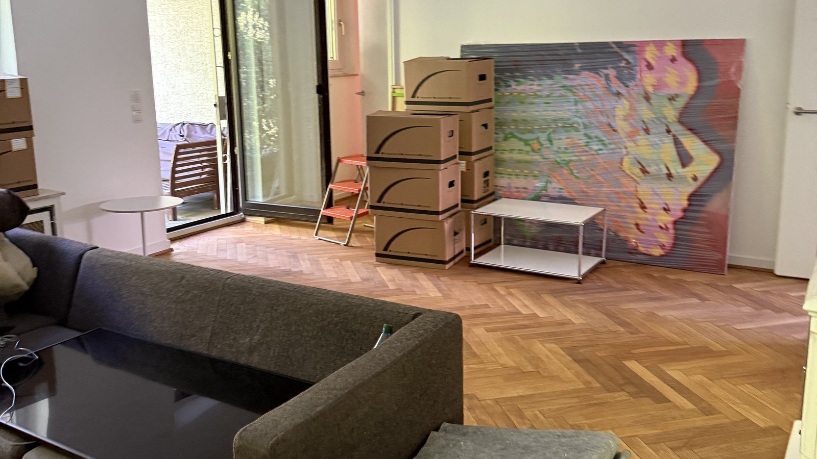 Umzugscheckliste für Ihren Umzug in Köln: Gestapelte Umzugskartons neben Sofa und Beistelltisch in einem Wohnzimmer mit Fischgrätparkett, an der Wand lehnt ein buntes Bild