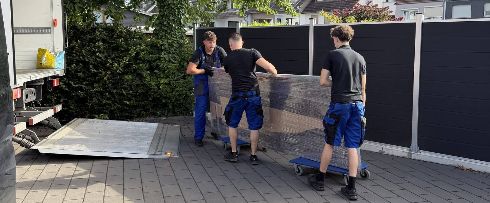 Umzugsunternehmen Köln: Drei Mitarbeiter schieben ein in Folie verpacktes Möbelstück auf Rollbrettern über den Hof zur Ladebordwand des Umzugswagens
