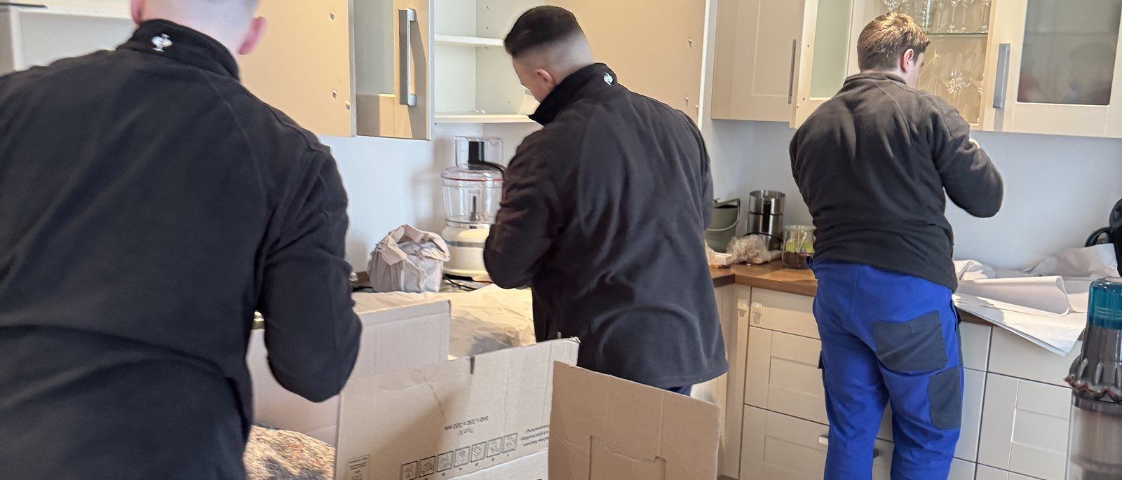 Verpackungsmaterial für Ihren Umzug in Köln: Drei Mitarbeiter packen in einer Küche mit hellen Fronten Geschirr und Kleingeräte in Umzugskartons auf der Arbeitsplatte